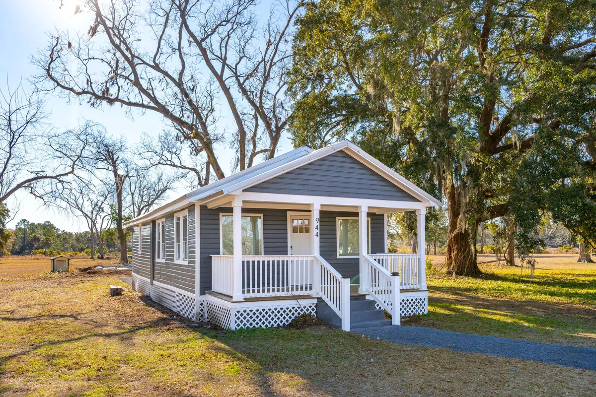 This image features the inviting front porch shaded by large trees, with the gray exterior of the house providing a serene backdrop. The porch is spacious and perfectly situated for relaxing, watching sunsets, or enjoying a quiet drink while soaking in the tranquil surroundings.