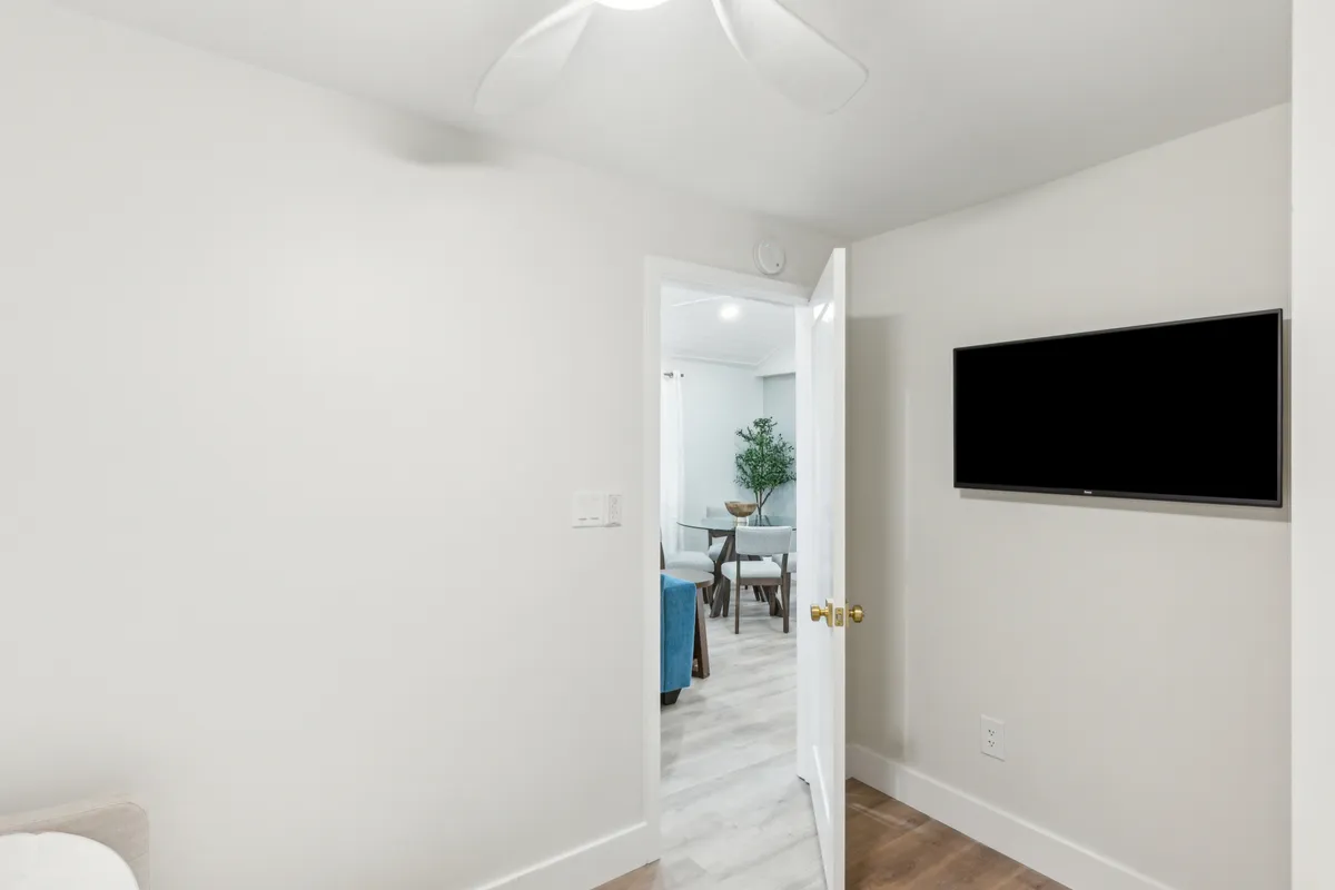 This image reveals a welcoming living area with an open door, ceiling fan, TV on the wall, a plant in the background, and a dining area visible through the doorway. The layout encourages relaxation and socializing, with sight-lines connecting the living and dining spaces, plenty of natural light, and touches of greenery for added warmth.
