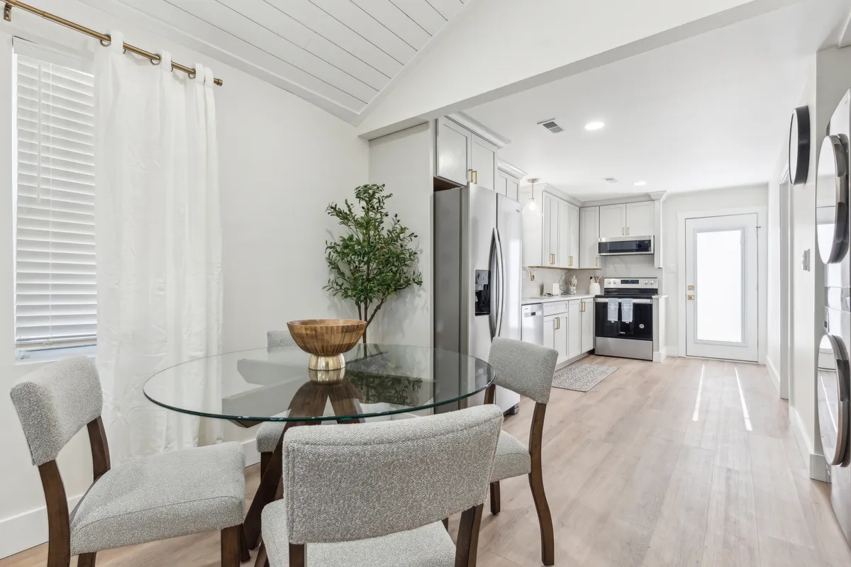 This bright interior scene focuses on a glass dining table set near the kitchen, surrounded by modern kitchen appliances and a potted plant. The atmosphere is cozy and sociable, ideal for group meals, celebrations, or a casual chat over coffee, with ample space for gathering.