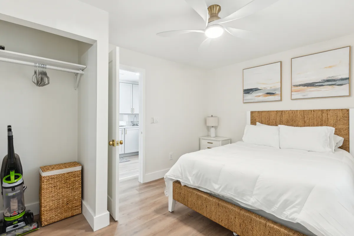 A bright, peaceful bedroom with a bed dressed in crisp white linens, a woven headboard, thoughtful wall art, and a nightstand with a soft-glowing lamp. Sunlight streams in, adding to the atmosphere of calm and restfulness.